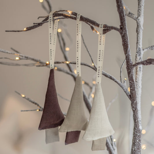 Three linen Christmas tree decorations hanging from a tree, in beige, brown, and white colorways, with branded hanging ribbons.