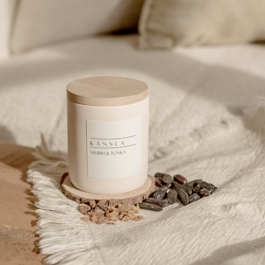 A concrete candle with a wooden lid, branded 'KANSLA', placed on a textured fabric, surrounded by some dried tonka beans.