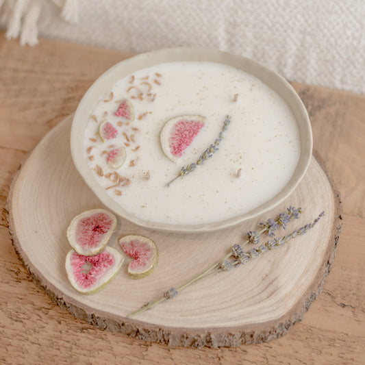 A limited edition centrepiece candle with wild fig, cassis, jasmine, and cedarwood scents, presented in a ceramic bowl with decorative dried fig, lavender, and orange blossom. The bowl is accompanied by decorative elements and sliced figs.