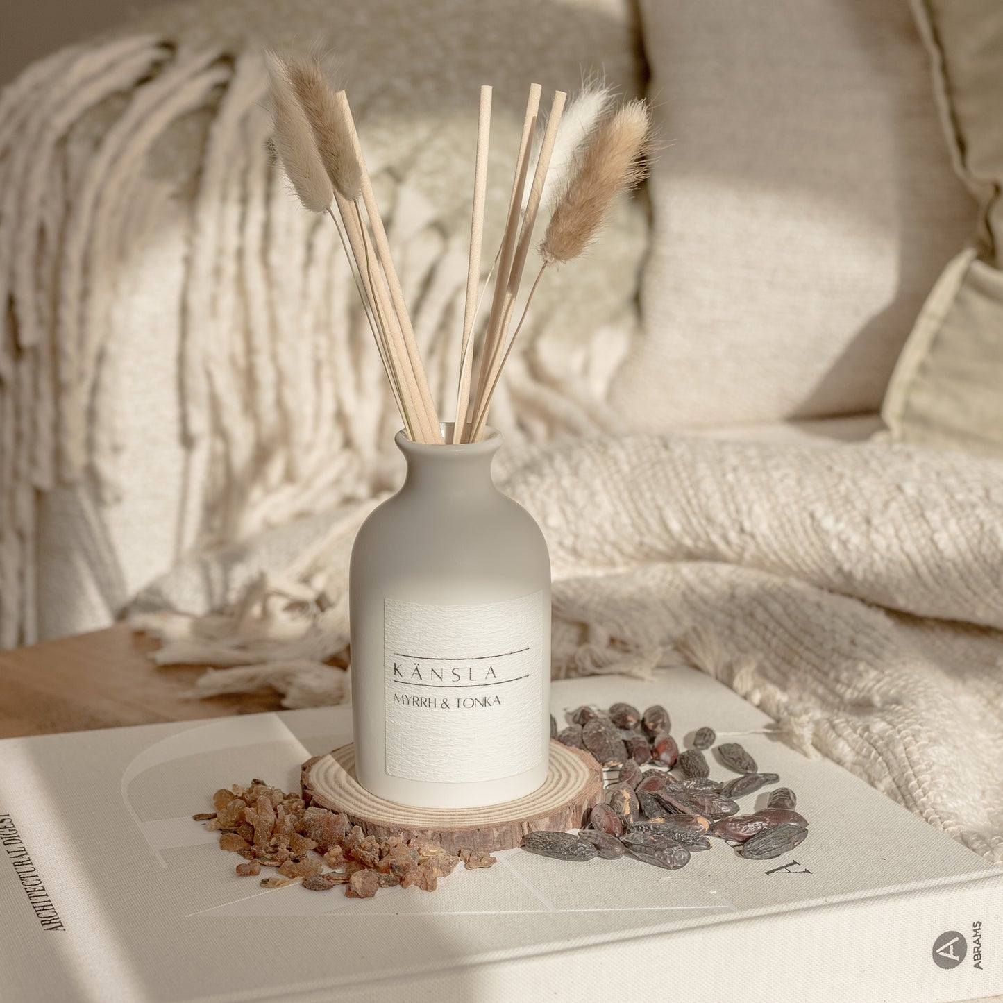 ceramic reed diffuser with natural rattan reeds and dried bunny tails. a white label reading kansla myrrh and tonka. placed on a decorative book with dried tonka beans around the sides