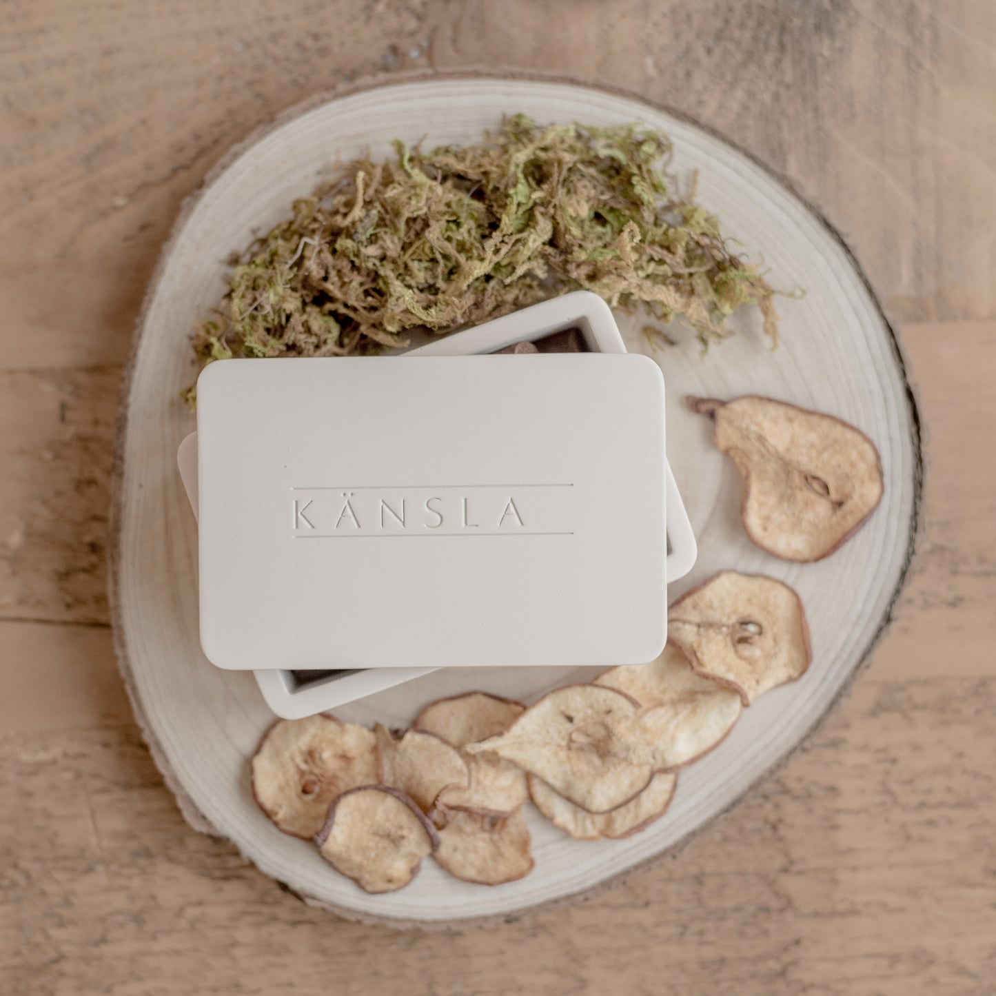 a birds eye view of a concrete box with an embossed kansla logo. the lid of the box is slightly off to one side to slightly show the incense cones inside. next to the box is decorative dried pear and moss