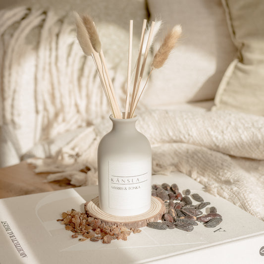 ceramic reed diffuser with natural rattan reeds and dried bunny tails. a white label reading kansla myrrh and tonka. placed on a decorative book with dried tonka beans around the sides