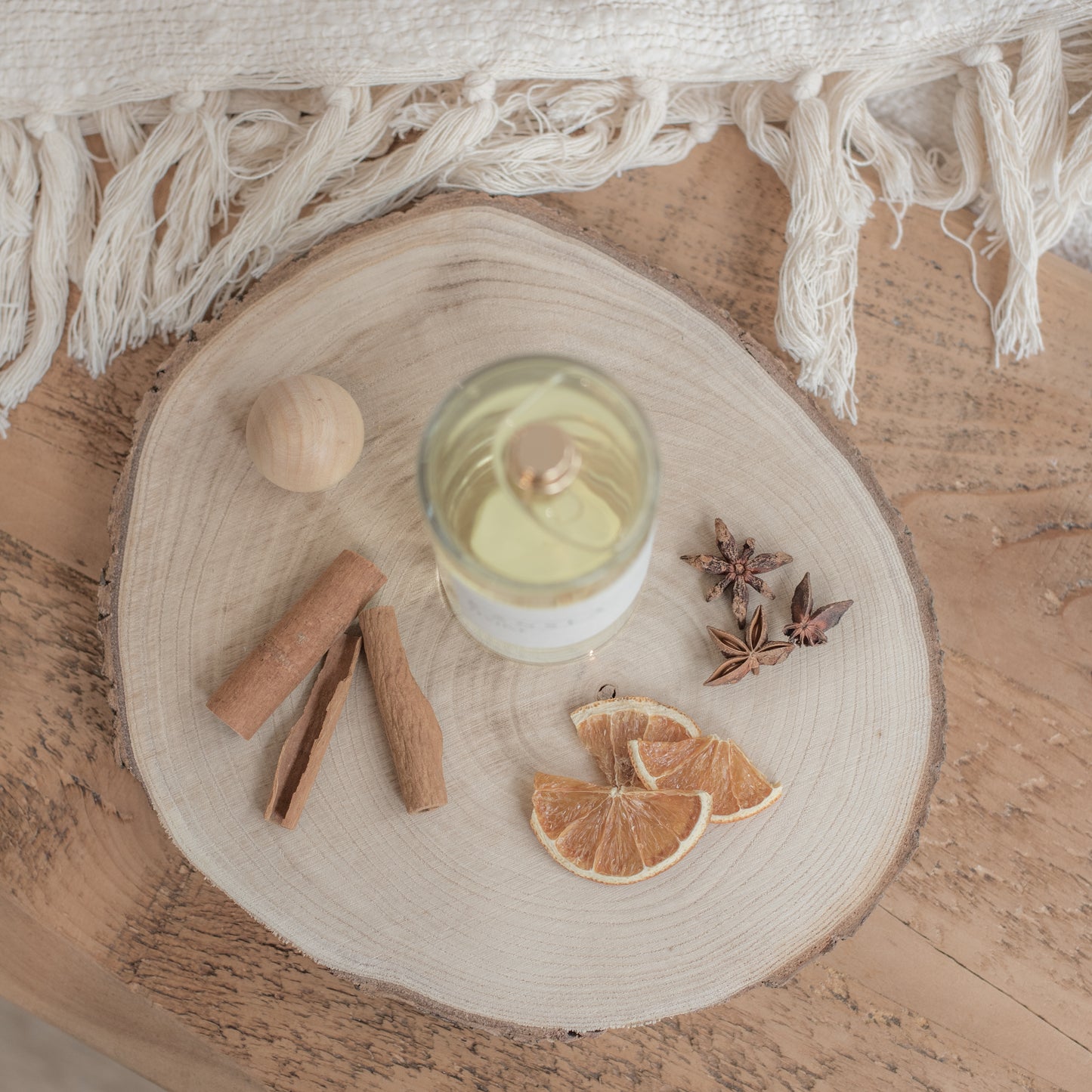 A picture taken from birds eye view of a clear glass bottle with a gold spray pump. The room spray is festive scent with dried orange and cinnamon decorations on a wooden tray