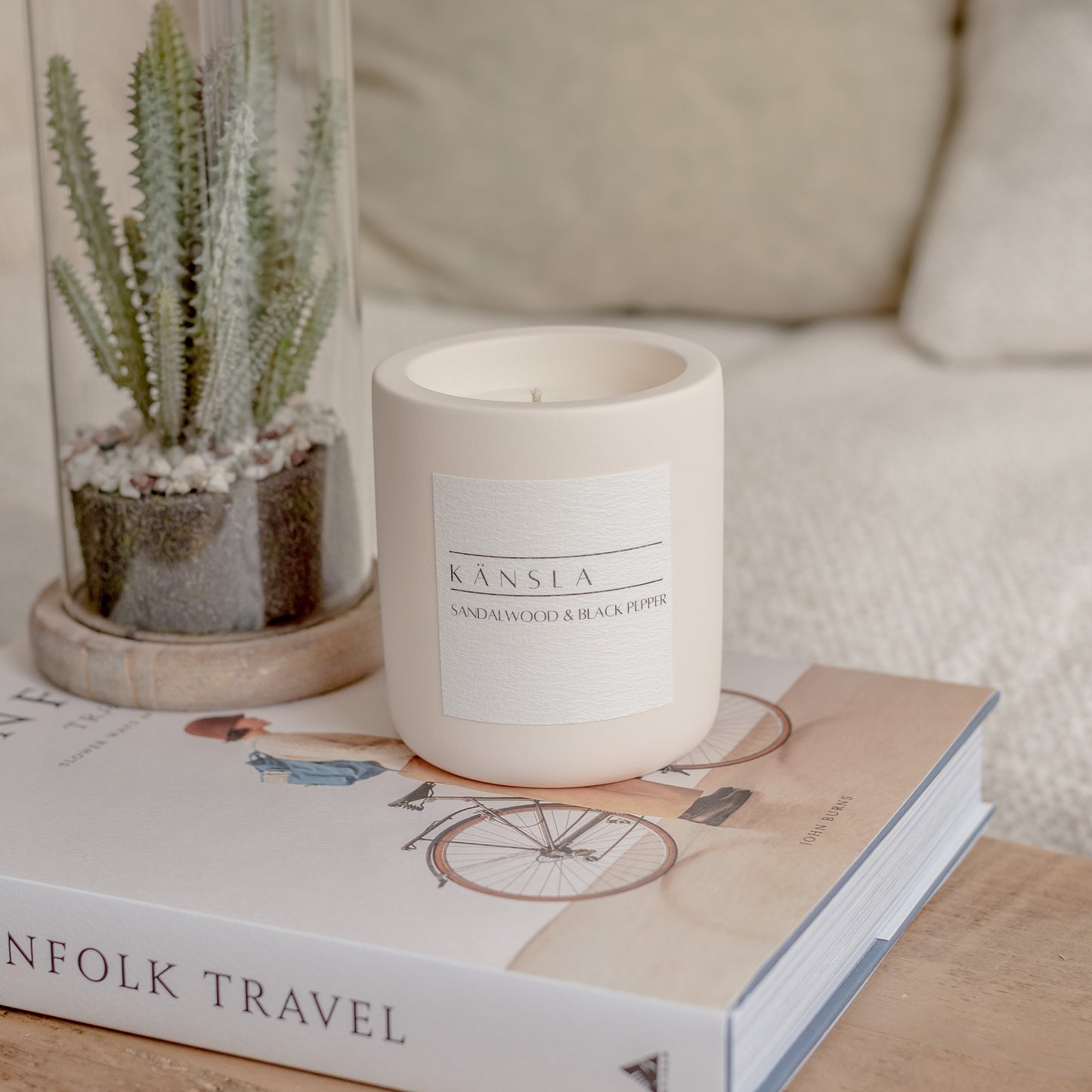 A concrete candle vessel w labelled 'KäNSLA SANDALWOOD & BLACK PEPPER', placed on top of a decorative styling book with a small plant in the background as decoration