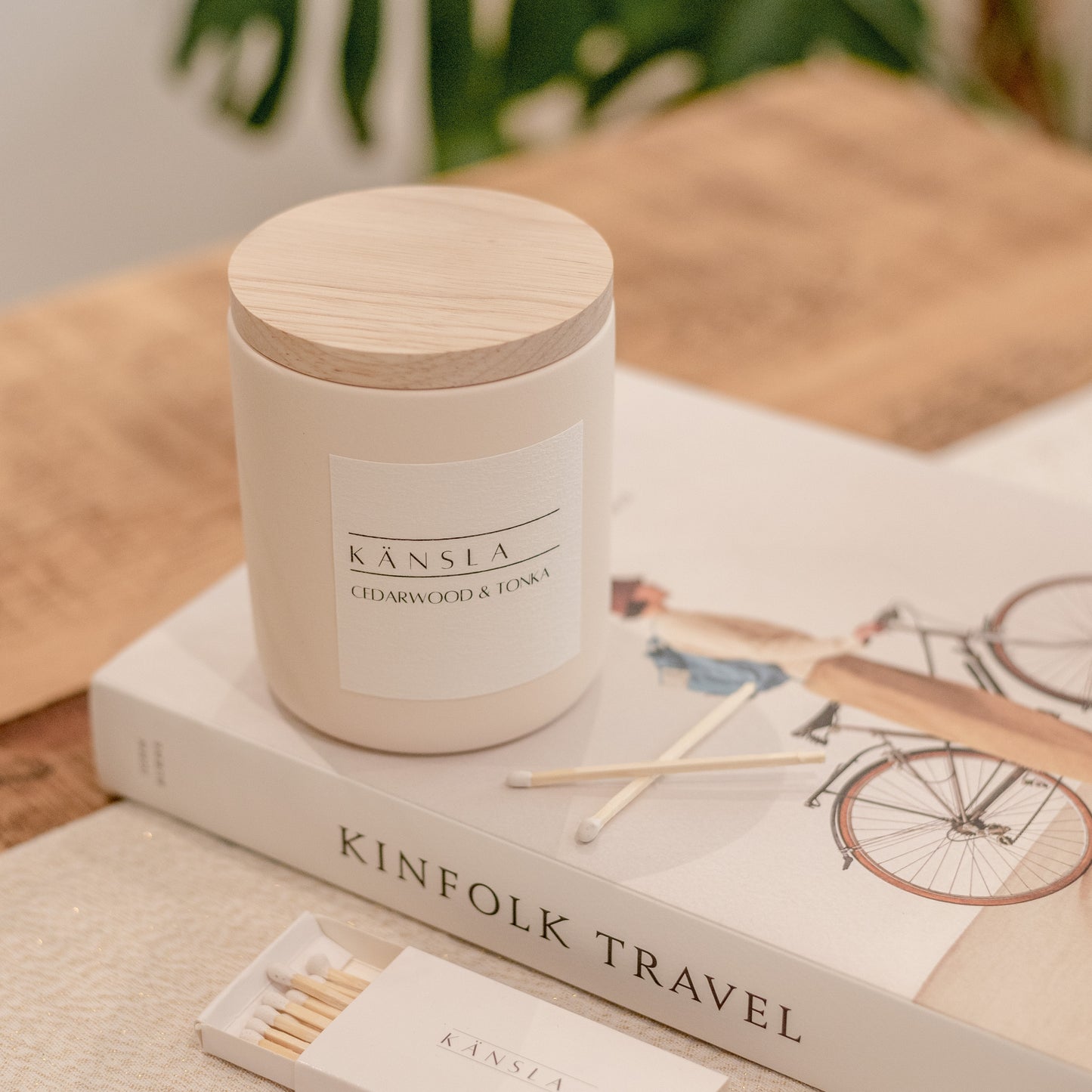 a high angle image of a concrete candle with a wooden lid. the candle has a white label on it that says kansla cedarwood and tonka. it is placed on a decorative book with 2 loose matches in front of it