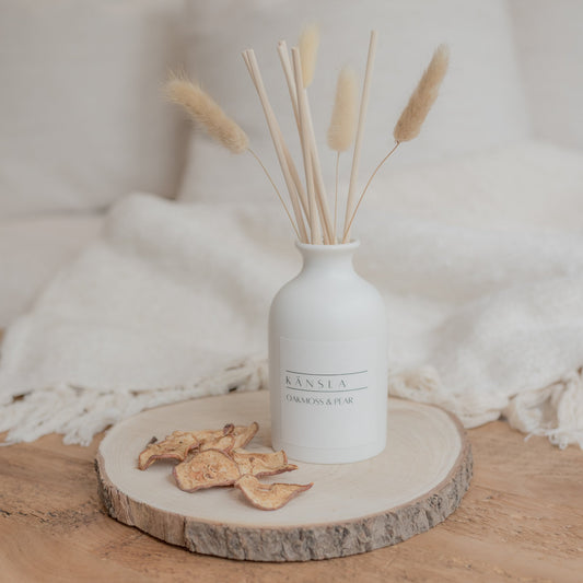 White ceramic reed diffuser with natural reeds and dried bunny tails oakmoss and pear scent. There is a white label on the bottle that says kansla oakmoss and pear