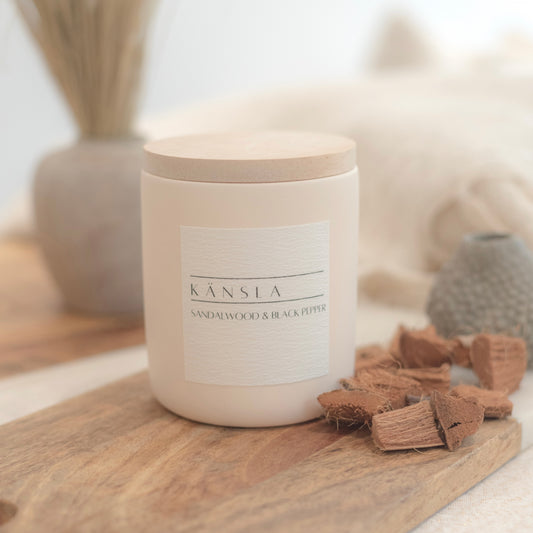 A beige concrete candle with wooden lid saying kansla sandalwood and black pepper. Placed on a wooden surface with decorative home styling in the background