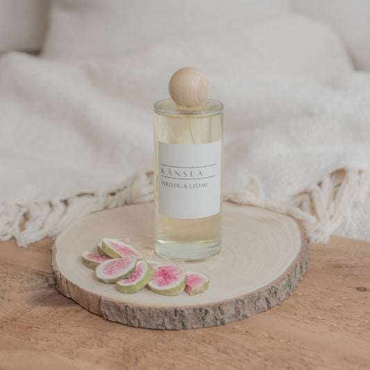 A luxury room perfume in a glass bottle with clear liquid inside. A wooden lid and a label that says kansla wild fig and cedar. It is placed on a wooden plate with dried decorative figs around the outside