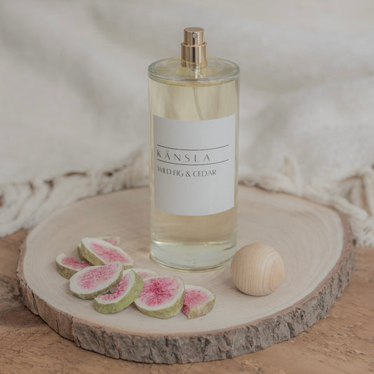 A luxurious room spray in a glass bottle with a gold spray pump. There is a wooden lid off to the side. This is a lifestyle shot placed on a wooden tray with soft furnishings in the background.
