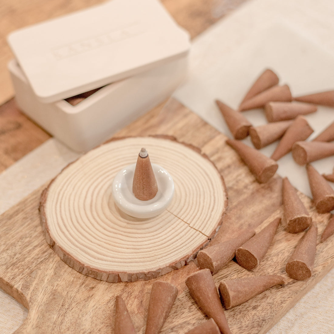 An image of a number of incense cones scattered on a wooden surface. There is a white ceramic burning dish with a lit incense cone in. Behind this is a kansla concrere incense box holder with the lid slightly off and the kansla embossed logo on the top 
