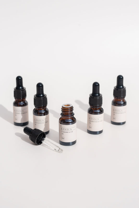 An image of 5 amber glass bottles with black lids containing oils. They are positioned in a v shape with the oil bottle at the front with no lid. There is a glass pipette with one oil in to the side all on a white background