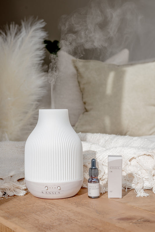 An image of a ceramic aroma diffuser with a white lid and rubber base. Mist is coming out the top and there is a small bottle and box of oil next to it