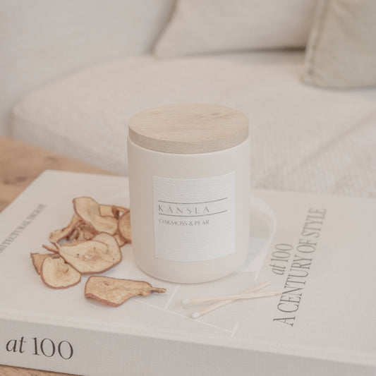 An image of a kansla Oakmoss and pear candle in a concrete jar and a wooden lid. It is placed on top of a styling book with dried pears around the side for decoration. In the background there is a soft beige sofa 