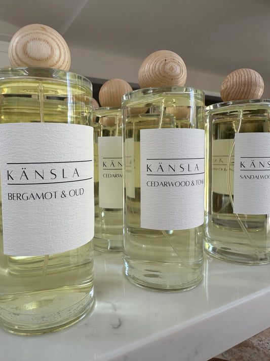 An image of multiple room perfumes in clear glass bottles with wooden lids. The room spray has clear liquid inside and a white label that has kansla branding and the scent names of the room perfumes 