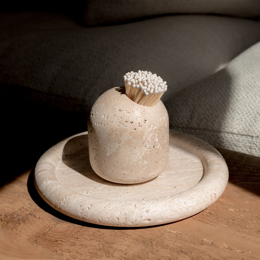 An image of a travertine match pot with white tipped matches in. The match pot is positioned on a travertine Tray on a wooden bench. There is a shadow in the background emphasising the focus on the match pot 