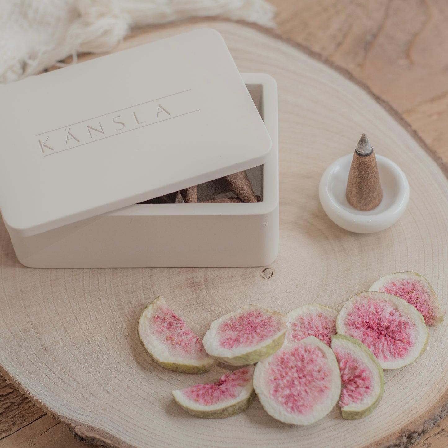 A close up shot of a concrete Kansla incense cone box with the lid slightly off. A white ceramic burning dish is to the side and there is a lit incense cone inside it. There are decorative dried figs in the foreground on top of a wooden plate