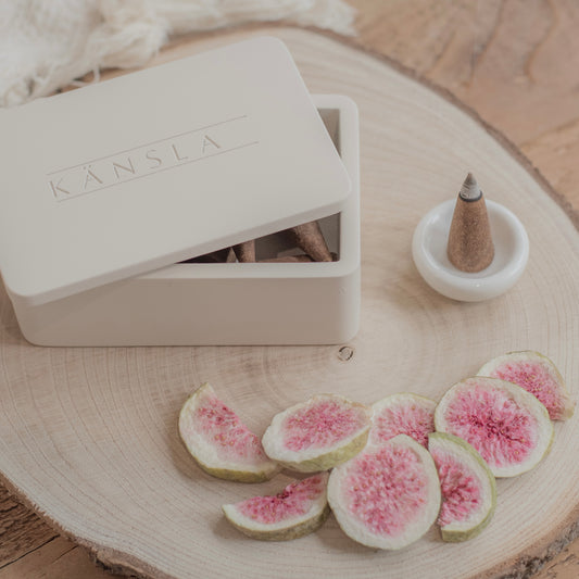 A close up shot of a concrete Kansla incense cone box with the lid slightly off. A white ceramic burning dish is to the side and there is a lit incense cone inside it. There are decorative dried figs in the foreground on top of a wooden plate
