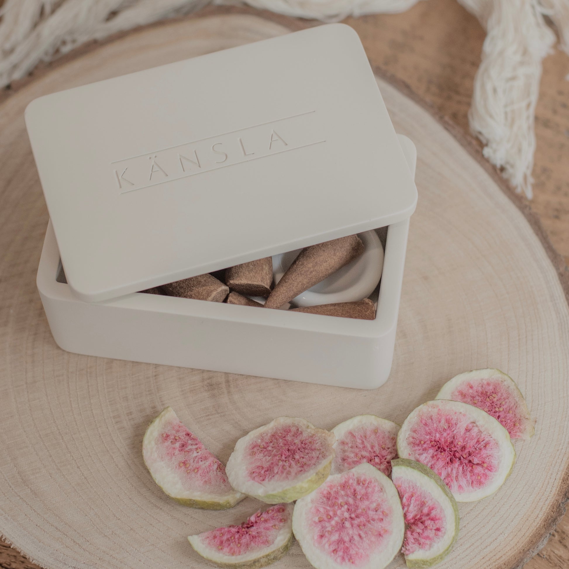 Close up of a concrete Kansla incense cone box with the lid slightly off. This is a lifestyle shot and there are dried decorative figs around the outside. The product is placed on a wooden bench with a soft throw in the background. You get a small glimpse of the incense cones inside the box