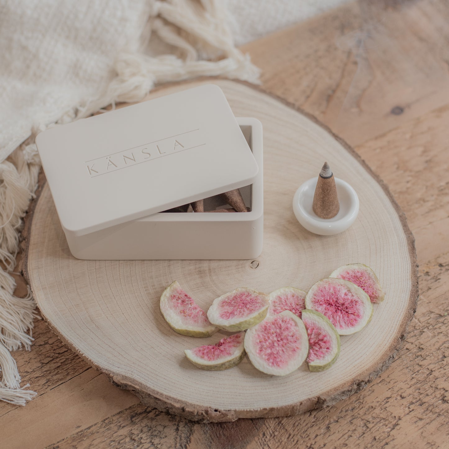 A concrete Kansla incense cone box with the lid slightly off. A white ceramic burning dish is to the side and there is a lit incense cone inside it. This is a lifestyle shot and there are dried decorative figs around the outside. The product is placed on a wooden bench with a soft throw in the background