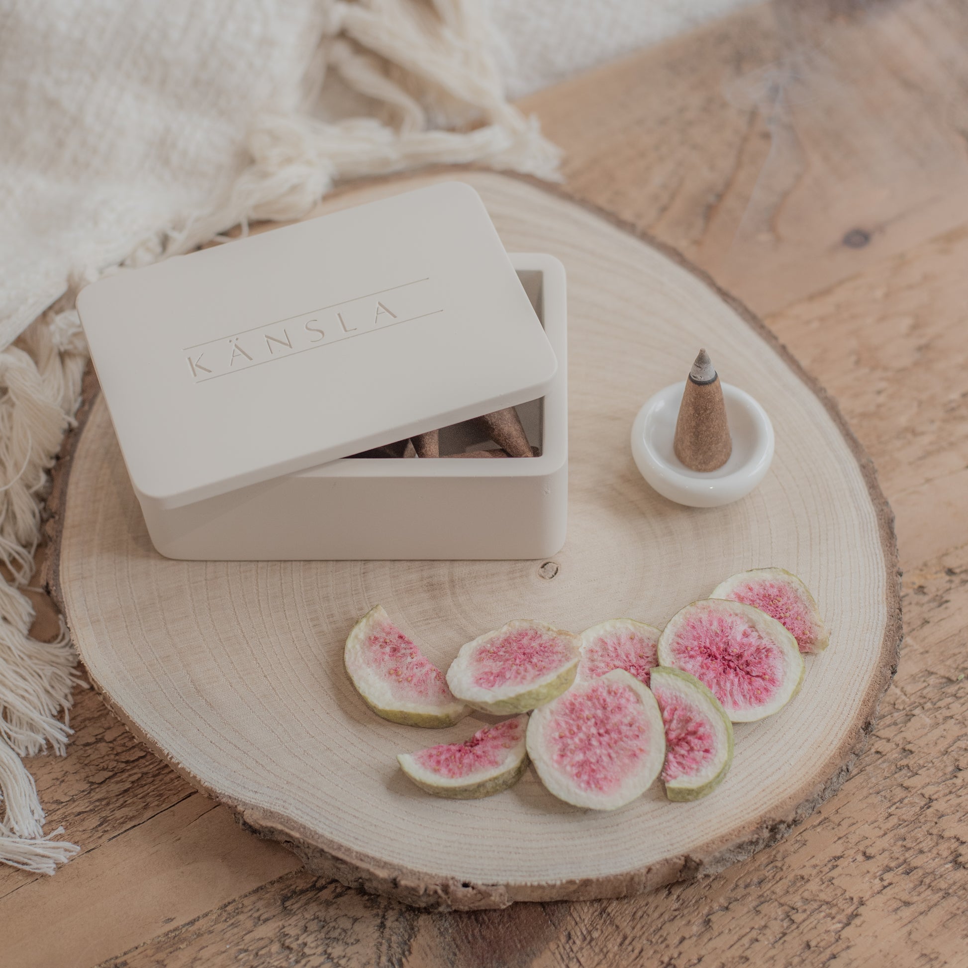 A concrete Kansla incense cone box with the lid slightly off. A white ceramic burning dish is to the side and there is a lit incense cone inside it. This is a lifestyle shot and there are dried decorative figs around the outside. The product is placed on a wooden bench with a soft throw in the background