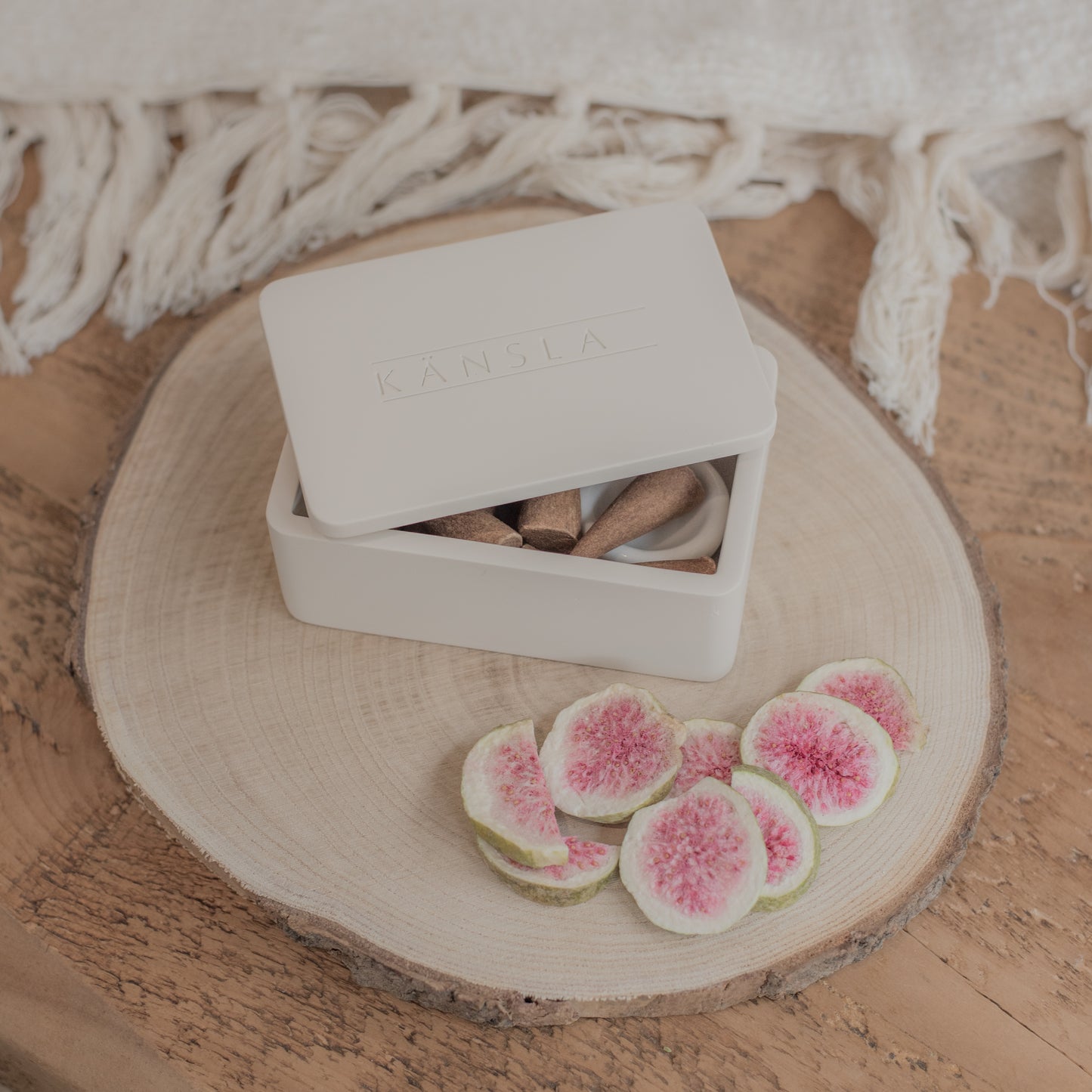 A lifestyle shot of a kansla incense cone box placed on a decorative wooden tray with a soft throw in the background and dried decorative gifs in the foreground. Inside the box you can see a glimpse of the incense cones and the ceramic burning dish