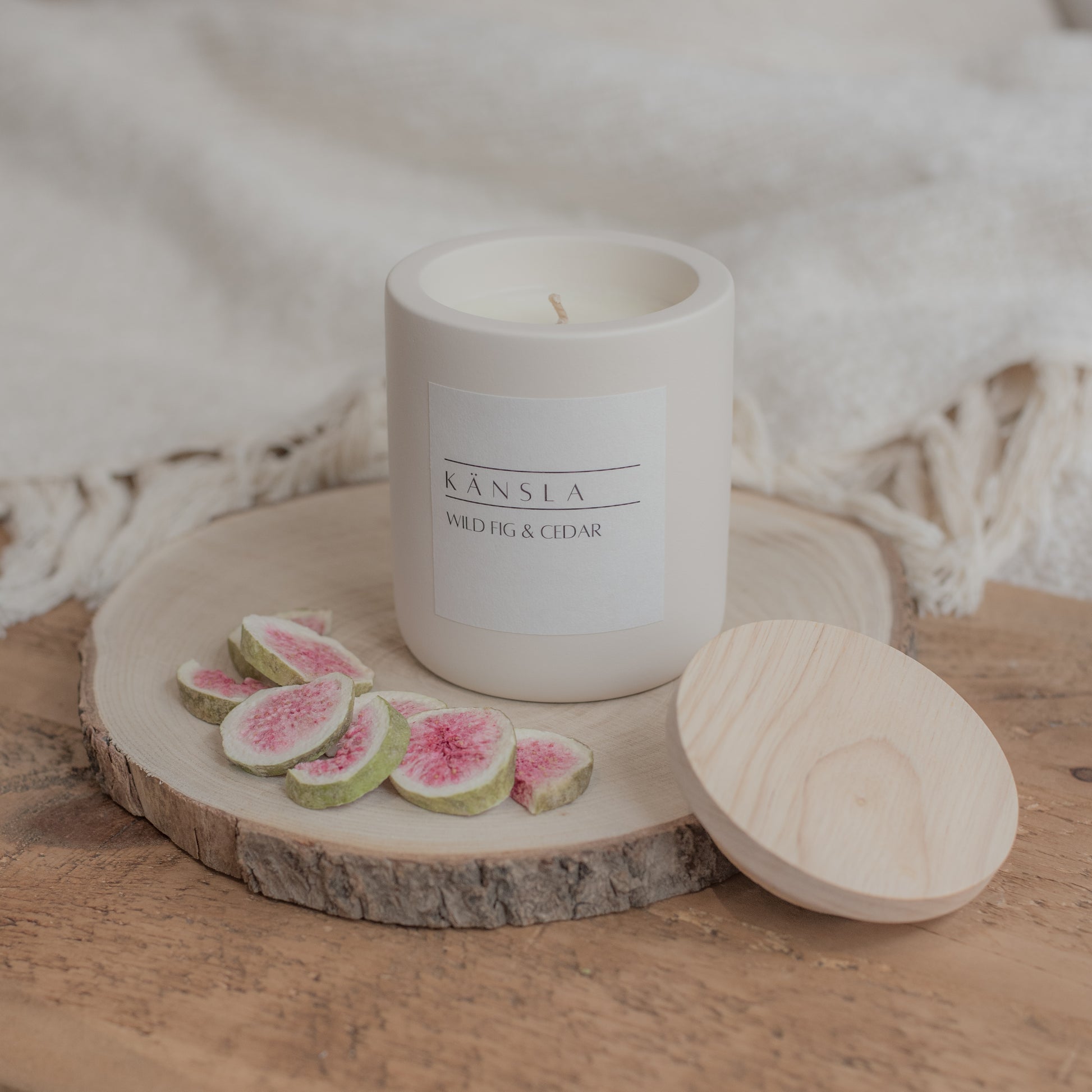 Candle with a wooden lid on a wooden surface with sliced figs. A lifestyle shot with soft furnishings in the background 