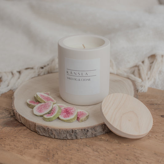 Candle with a wooden lid on a wooden surface with sliced figs. A lifestyle shot with soft furnishings in the background 