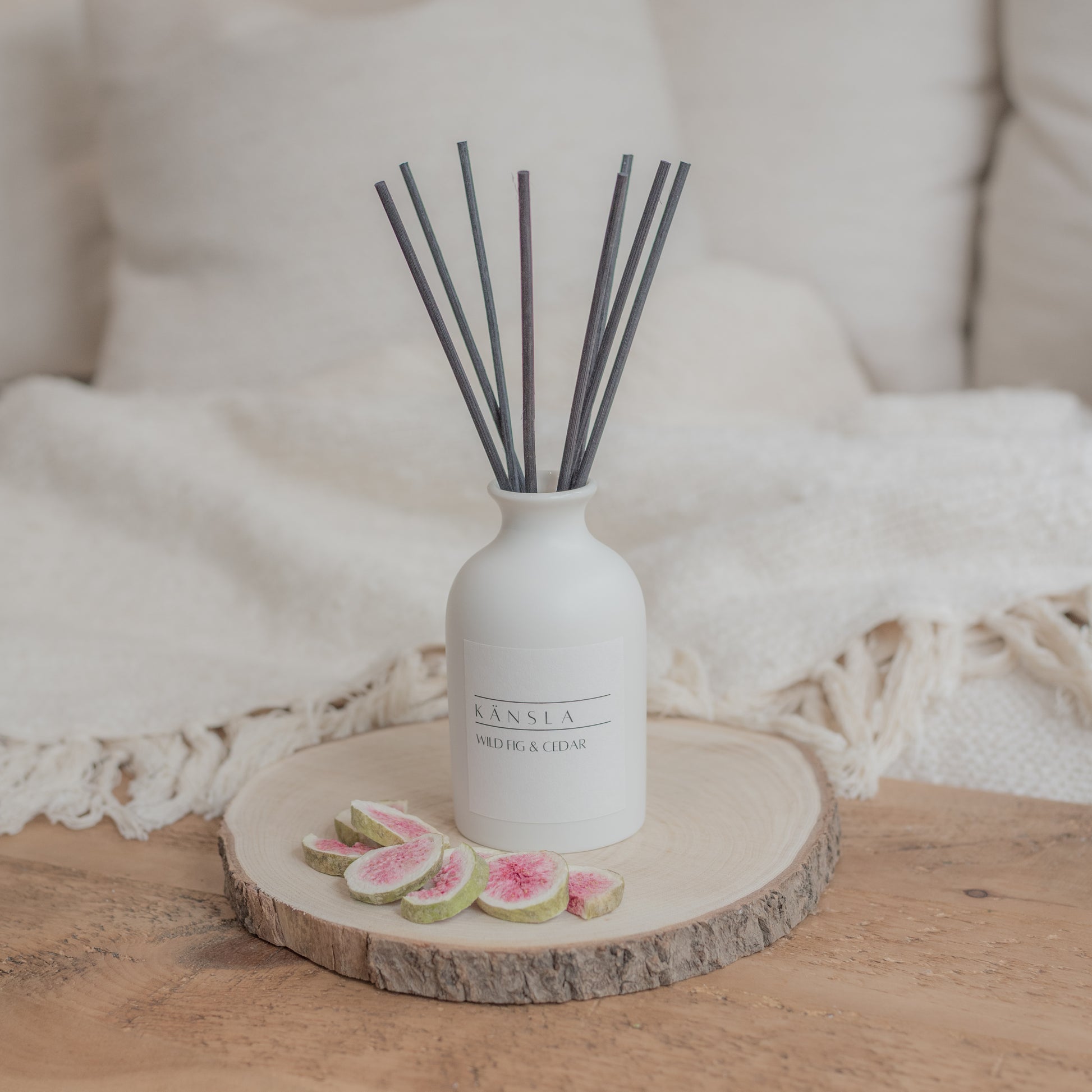 White ceramic reed diffuser on a wooden plate with black rattan reeds. The background has soft furnishings setting the scene 