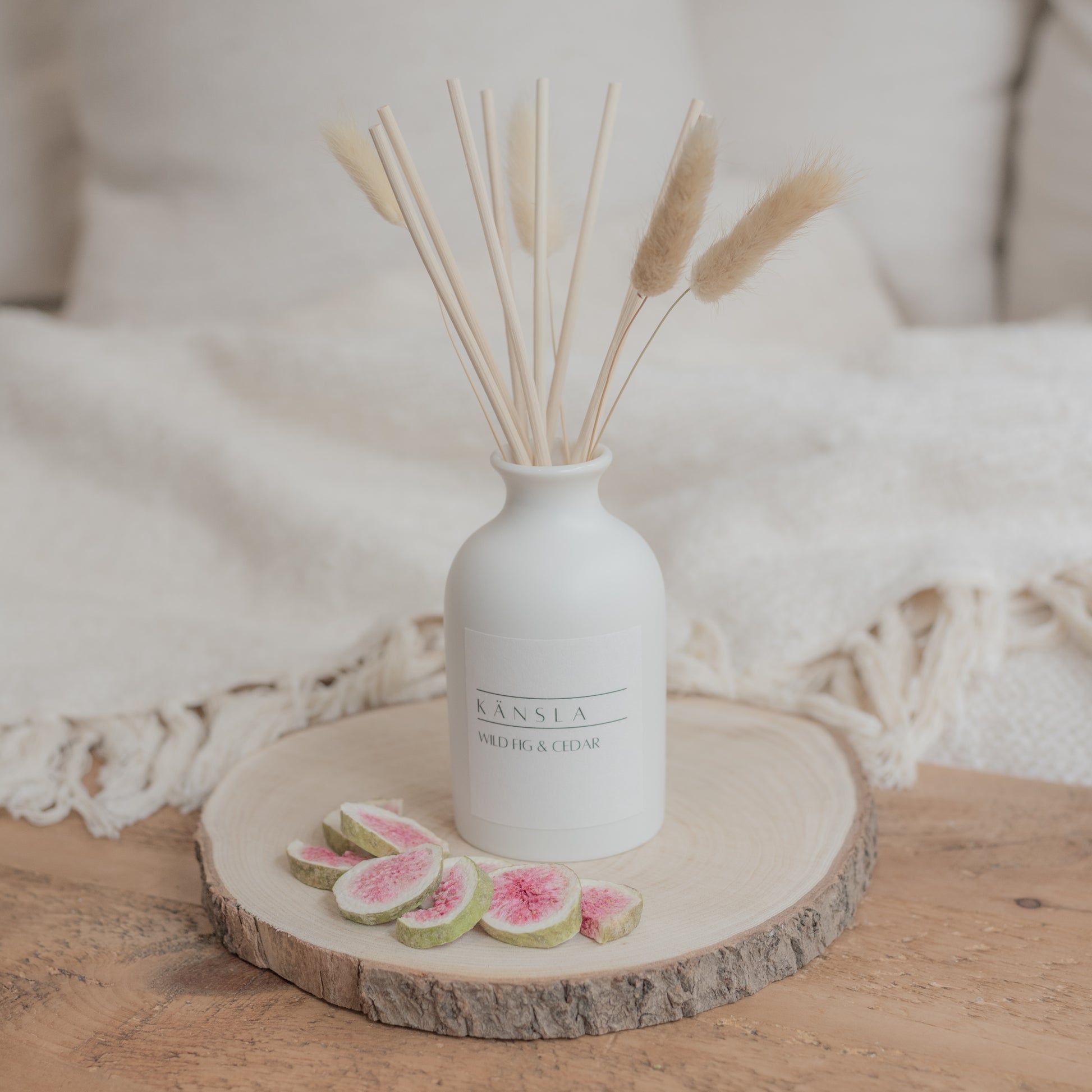 A kansla reed diffuser in a lifestyle shot. Placed on a wooden bench with dried fig around the outside. The reed diffuser is matte white ceramic and there is a white label with black writing saying wild fig and cedar kansla. There are natural rattan reeds nd dried decorative bunny tails inside 