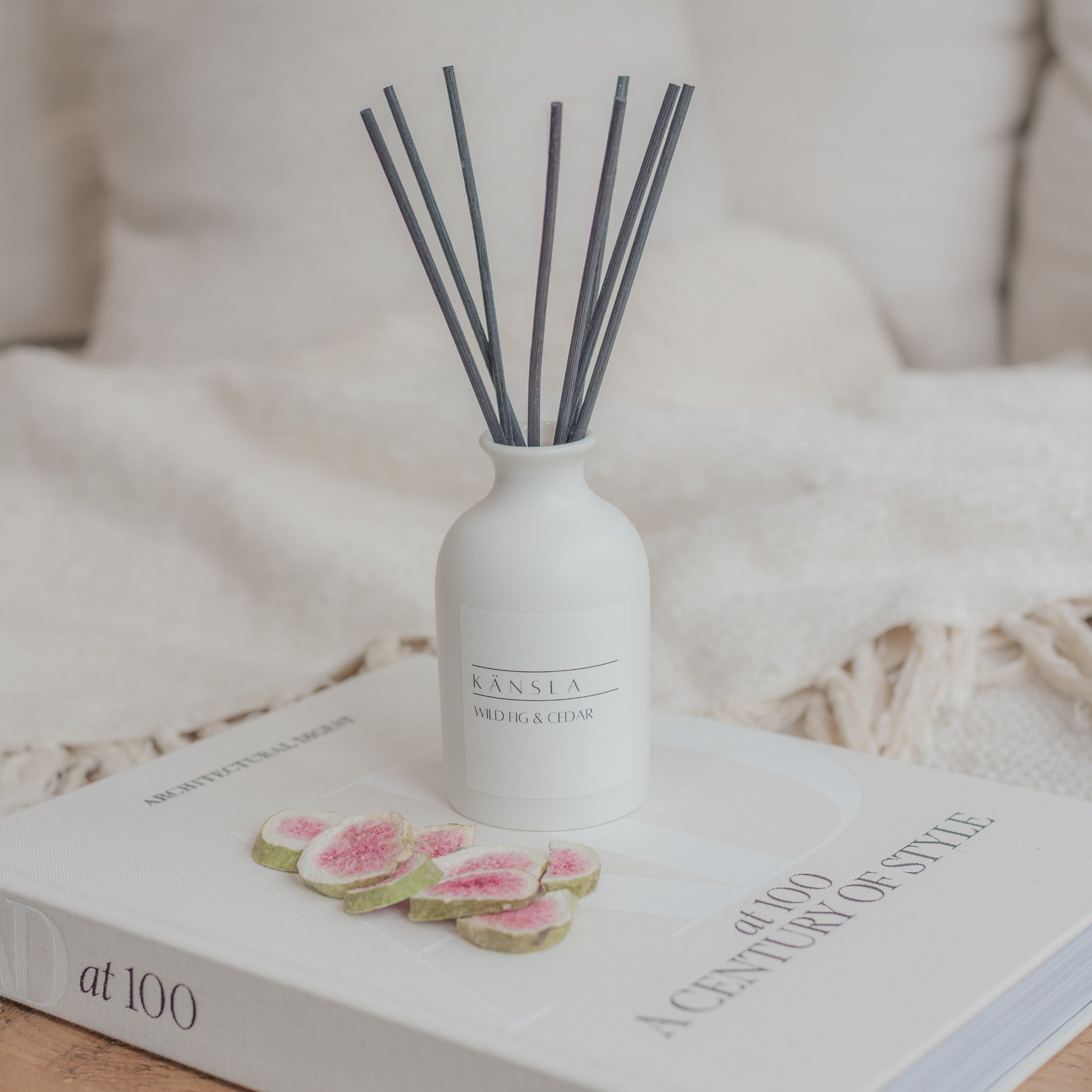 A lifestyle shot of Känsla’s wild fig and cedar reed diffuser. Placed on a decorative book the diffuser has black rattan reeds and a white ceramic jar with a white label on the front reading the scent name 