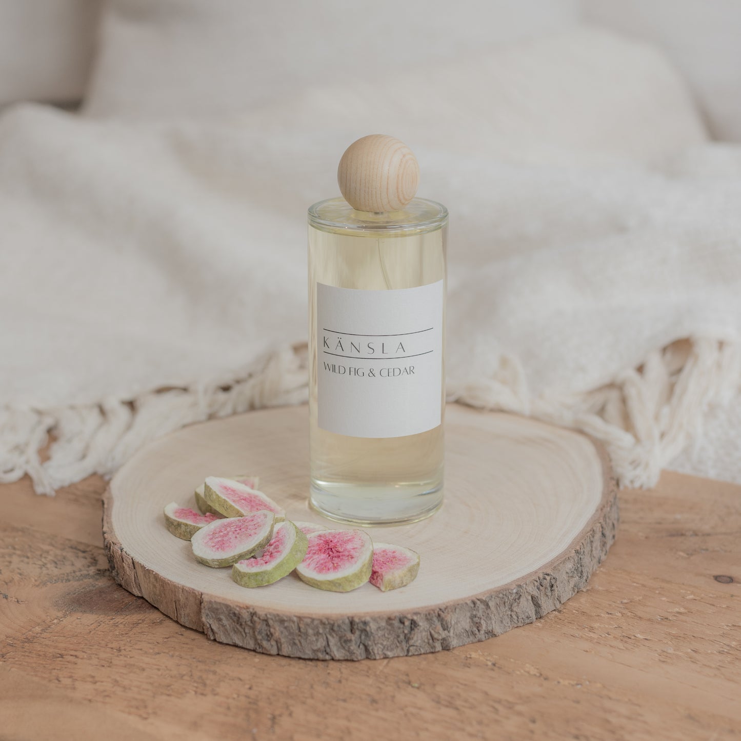 A luxury room perfume in a glass bottle with clear liquid inside. A wooden lid and a label that says kansla wild fig and cedar. It is placed on a wooden plate with dried decorative figs around the outside 