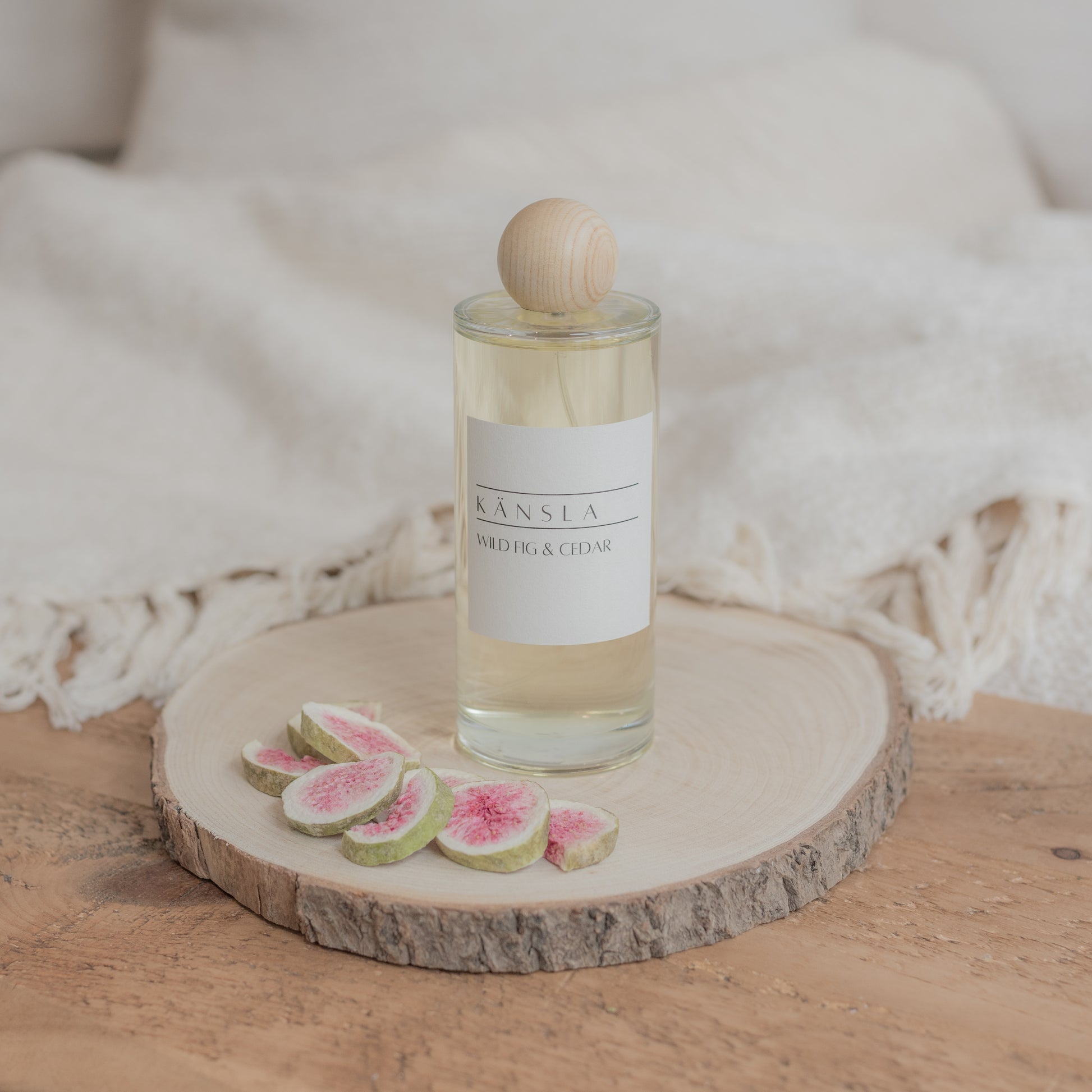 A luxury room perfume in a glass bottle with clear liquid inside. A wooden lid and a label that says kansla wild fig and cedar. It is placed on a wooden plate with dried decorative figs around the outside 