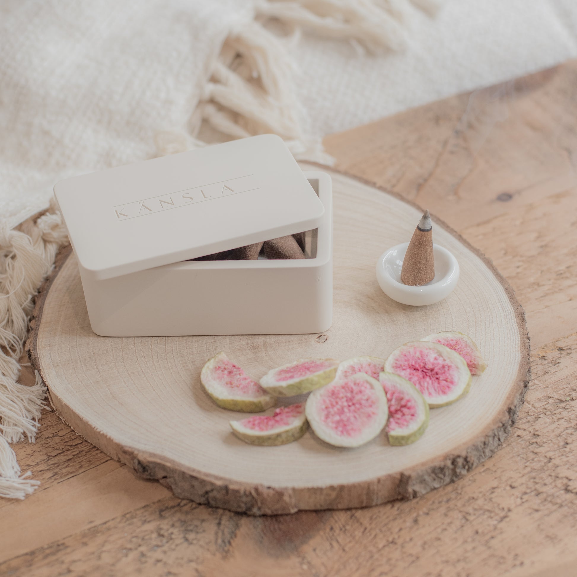 A lifestyle shot on a wooden bench with a soft throw in the background. the foreground shows a Kansla concrete incense cone box with a lit cone burning next to it in a ceramic burning dish, finished with decorative dried figs in the foreground